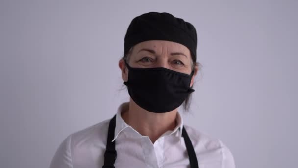 Uzavřené obchody během uzamčení. Portrét kuchařky s naběračkou v rukou na šedém pozadí. Šéfkuchař nosí černou masku, klobouk, rukavice a zástěru. — Stock video