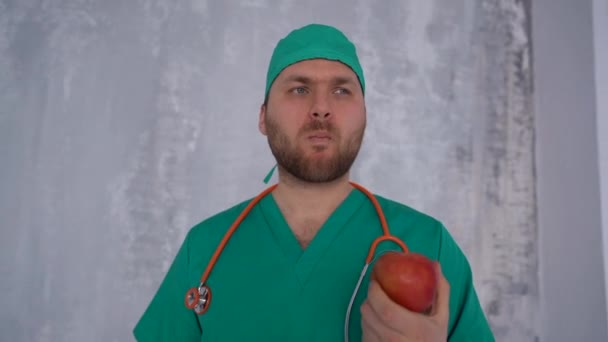 Młody lekarz w zielonym garniturze i czapce z fondoskopem na ramionach jedzący jabłko — Wideo stockowe