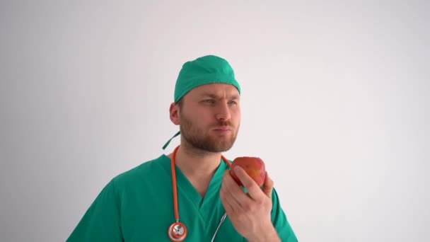 Fiatal férfi szakállas orvos terapeuta eszik egy almát munka közben. Egészséges táplálkozás, orvosi szakma — Stock videók