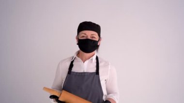 Yeni gerçeklik. Üniformalı, koruyucu maskeli ve siyah eldivenli kadın bir aşçının portresi. Tahta bir oklava tutan bir kadın.
