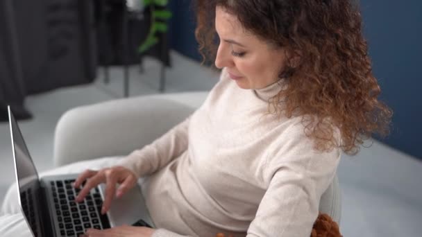 Vista dall'alto di una donna freelance che lavora a casa, digitando sulla tastiera. Ritratto di giovane donna riccia seduta su una sedia con un computer portatile e un plaid. Lavoro a distanza, concetto di home office — Video Stock