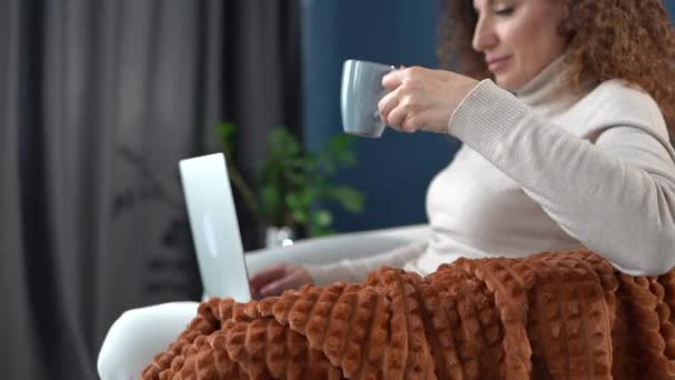 Blijf thuis concept. Meisje drinkt koffie en werkt met behulp van een laptop tijdens het zitten in een gezellige fauteuil thuis — Stockvideo