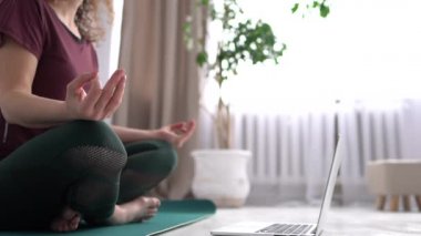Yetişkin bir kadın evde çalışıyor, yeşil minder üzerinde yoga egzersizi yapıyor, lotus pozisyonunda oturuyor ve meditasyon yapıyor, nefes alıyor, dinleniyor. Vücut yakın plan