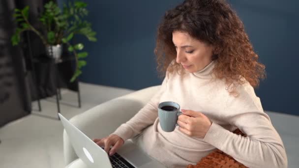 Retrato de una joven que trabaja usando un portátil. Una mujer bebe café y trabaja remotamente mientras está sentada en un cómodo sillón. Concepto de oficina — Vídeo de stock