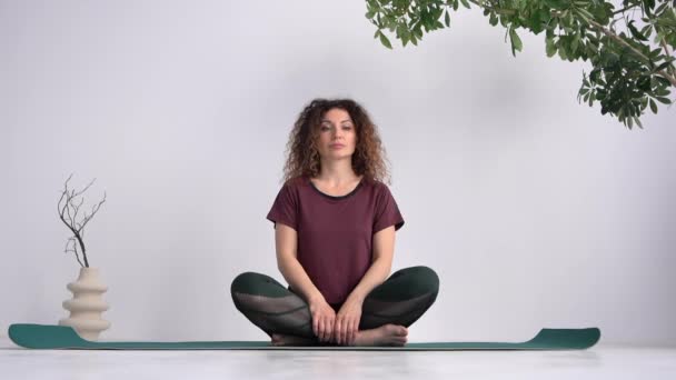 Lovely curly haired woman with serious focused face sits on mat in lotus position in yoga studio — Vídeos de Stock