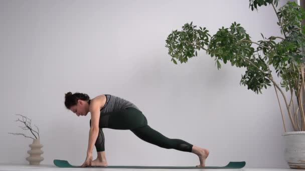 Adult woman in a sports top and leggings practices yoga on a mat. Mindfulness and wellbeing concept. In the frame there is a large indoor plant in a pot — Αρχείο Βίντεο