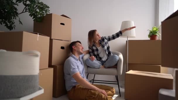 Happy young family discusses a plan for a new home immediately after relocation. Husband and wife sit among cardboard boxes — Stock Video