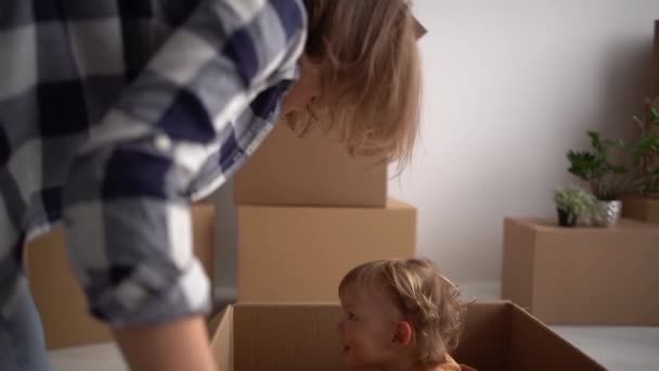 У мамы и сына есть фанат во время переезда. Молодая мама играет с ребенком, сидящим в картонной коробке. Семья переезжает в новую квартиру, концепция недвижимости — стоковое видео