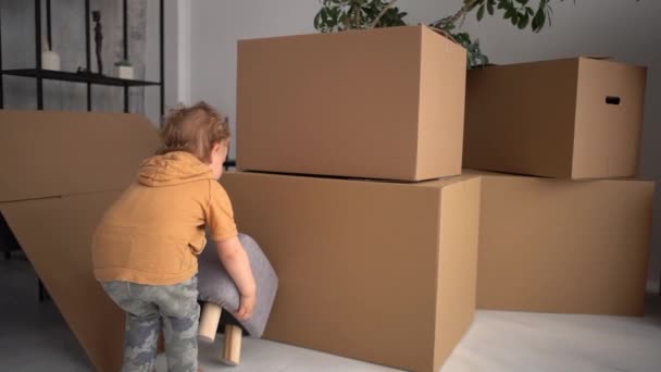 Peuter jongen draagt een kleine poef tegen de achtergrond van kartonnen dozen. Het kind helpt de ouders met de verhuizing naar een nieuw huis. Vastgoedconcept — Stockvideo
