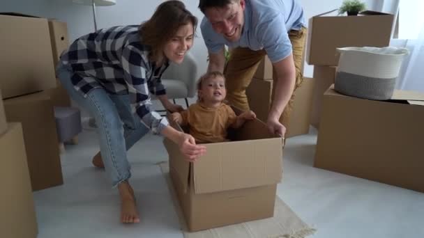 Lycklig familj, far, mor och son firar flytten till en ny lägenhet. Föräldrarna rullar barnet i lådan. Begreppet omlokalisering — Stockvideo