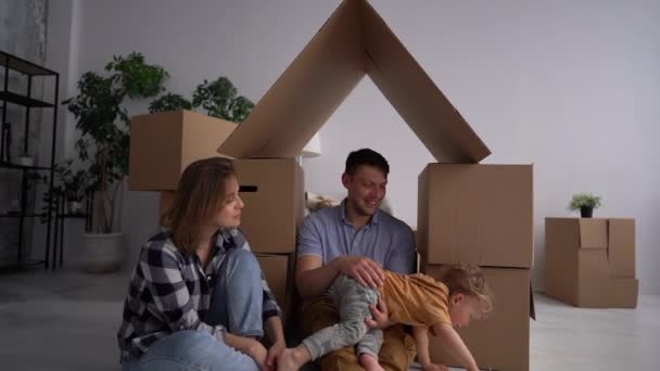 Família caucasiana, homem, mulher e menino criança sentar no chão, jogar e descompactar caixas e sorrir em nova casa. Atrás deles caixas móveis — Vídeo de Stock