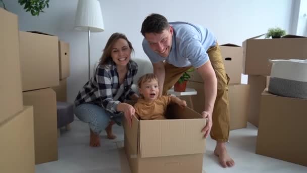 Ett lyckligt barn och hans unga föräldrar har roligt i sitt nya hem under flytten. Mamma och pappa rullar en liten pojke i en pappkartong på golvet — Stockvideo