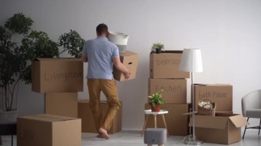 Yetişkin adam kutuların arkasında bir şeylerle dans ediyor. Adamın yeni bir eve taşınma zevki var. Mukavva kutular. Mutfak, oturma odası, garaj, banyo.