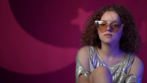 Retrato de una encantadora chica rizada con gafas elegantes en luces de neón en un club de discoteca. La adolescente lleva un vestido brillante. Estudio de rodaje — Vídeos de Stock