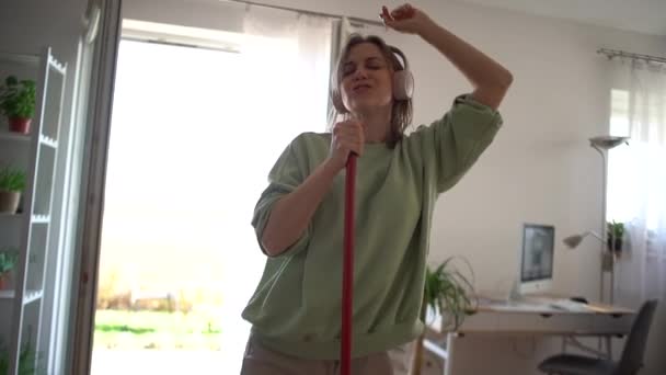 Hübsche junge Hausfrau wäscht den Boden in Loft-Stil Wohnung und hört Musik über Kopfhörer, sie singt und tanzt Spaß mit Wischmopp — Stockvideo