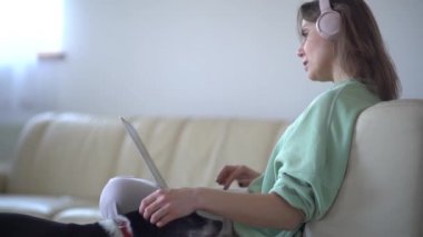 Evdeki kanepede oturan, kablosuz kulaklıkla müzik dinleyen ve dizüstü bilgisayar kullanan güzel bir yetişkin kadın. Kulaklıklı kız film izliyor, köpeğini okşuyor. Evde kal konsepti