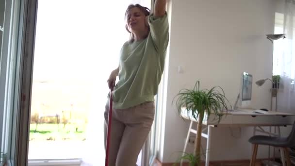 Mujer divirtiéndose durante la limpieza cantando y bailando con fregona disfrutando de la música en los auriculares. Hermosa ama de casa haciendo la tarea, limpieza de la casa, agencia de limpieza — Vídeos de Stock