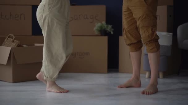 Close-up of the bare feet of an unrecognizable husband and wife against the background of cardboard boxes with things. Moving to a new house or apartment, real estate concept — Stock Video