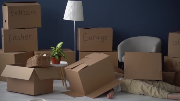 Adult woman lying on the floor under empty cardboard boxes against the background of boxes filled with things. Moving difficulties, decluttering and minimalism concept. Funny video — Stock Video