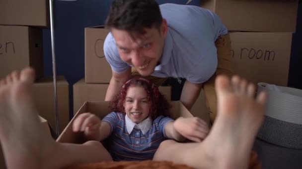 Близкий портрет девочки-подростка с босыми ногами сидит в картонной коробке, в то время как ее счастливый отец катает ее по новому дому — стоковое видео