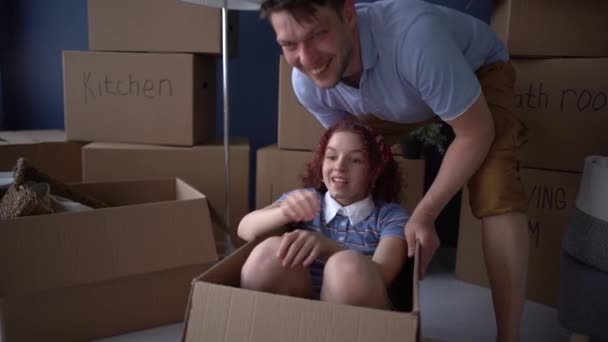 男は十代の娘を段ボール箱に転がします。幸せな父と娘は彼らの新しい家に移動の完了を祝う — ストック動画