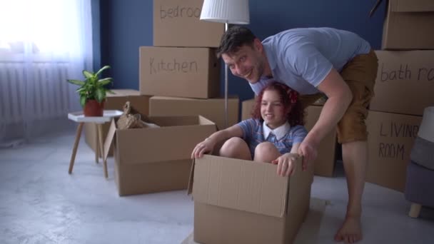 Buon padre e figlia celebrano il completamento del trasloco nella loro nuova casa. Uomo rotola sua figlia adolescente in una scatola di cartone — Video Stock