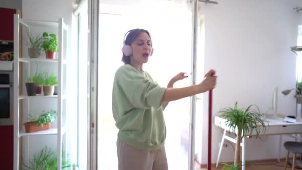 Mujer joven haciendo limpieza de la casa mientras baila. Encantadora ama de casa fregona el suelo y disfruta de la música con auriculares inalámbricos — Vídeos de Stock