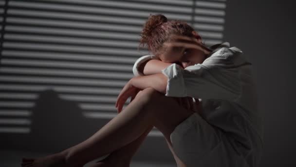 Close up of a sad girl in a white shirt sitting on the windowsill. Portrait of a teenage girl illuminated by evening light through the blinds. Stay at home concept — Stock Video