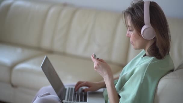 Retrato de uma jovem mulher em fones de ouvido com um cartão bancário em suas mãos e um laptop. Compras on-line, ficar em casa conceito — Vídeo de Stock