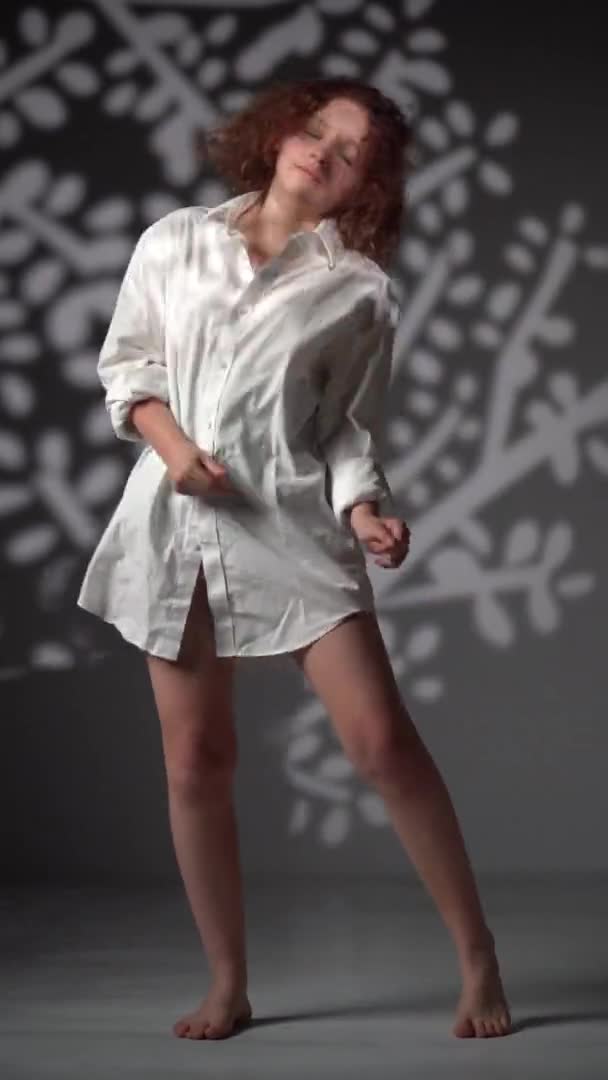 一个穿着白衬衫，赤脚站在白光背景下的少女的垂直视频肖像。圣诞晚会，创意拍摄理念 — 图库视频影像