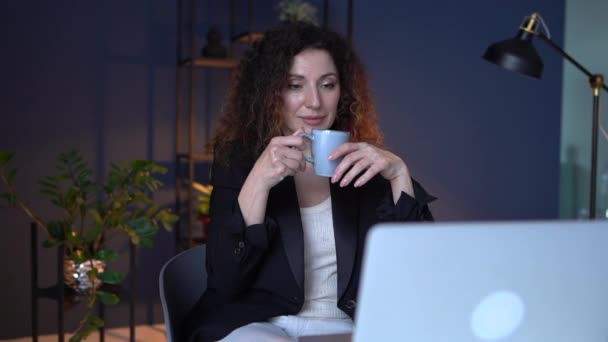 Empresaria bebe té mientras usa un portátil. Una mujer atractiva que trabaja hasta tarde en casa. Empleo remoto, concepto de oficina en casa — Vídeos de Stock