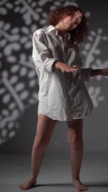 Dikey video portresi. Evde Noel partisi var. Çıplak ayaklı genç bir kız beyaz bir tişörtle arka planda beyaz bir ağaç şeklinde ışık ve gölgelerle dans ediyor.
