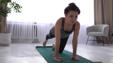 Güzel esmer kadın evde yoga minderinin üzerinde çalışıyor. Kız bacak ve sırt kasları için egzersizler yapıyor. Evde spor yapma etkinliği