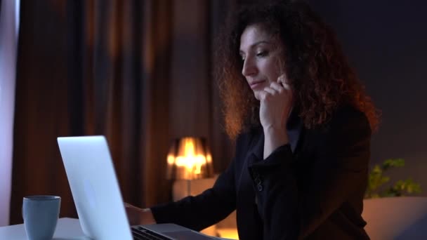 Hermosa mujer de negocios rizado en un traje trabaja mientras está sentado en una mesa con un ordenador portátil en una acogedora oficina o espacio de coworking. Una jefa exitosa y segura. Empleo remoto — Vídeos de Stock