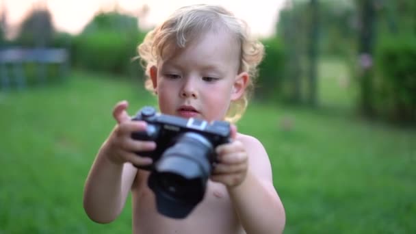 Zbliżenie portret uroczego blondyna z kamerą w rękach. Dziecko bada aparat podczas spaceru po podwórku w wiosce. Mały fotograf. — Wideo stockowe