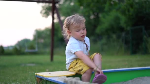 Chlapec sedí na pískovišti a sundává si boty. Dítě si v létě hraje na hřišti. Šťastné dětství sladké batole — Stock video