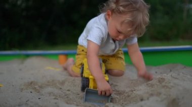 Küçük hevesli çocuk plastik bir çöp kamyonuyla oynuyor. Çocuk plastik bir oyuncakla kum havuzunda kum kazıyor.