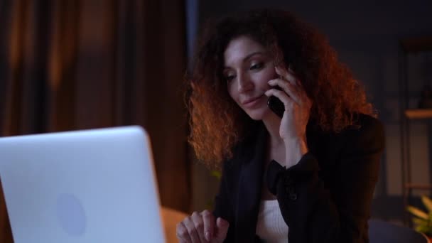 Atractiva mujer de negocios de pelo rizado es lleva a cabo las negociaciones de trabajo por teléfono mientras está sentado en la computadora tarde en la noche. Chica con exceso de trabajo trabajando en la oficina hasta tarde en la noche — Vídeos de Stock