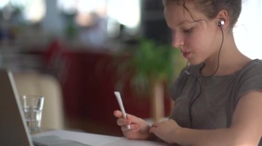 İnternette video dersi alan sevimli bir genç kızın portresi. Bir kız dizüstü bilgisayarın yanında oturuyor ve kulaklık takıyor, dinliyor ve yazıyor. Mutlu genç kız İngilizce öğreniyor.