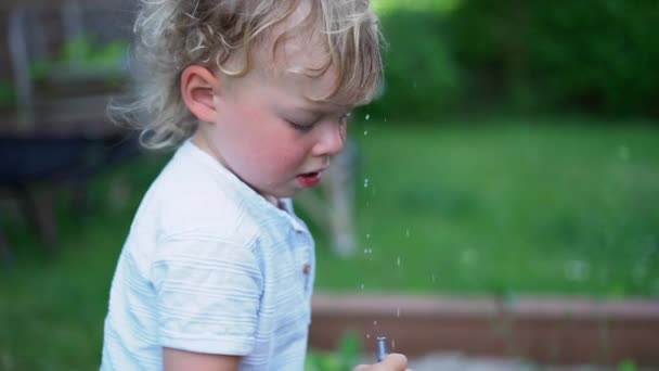 Retrato cercano de un rubio rizado jugando con una manguera en un huerto. Niño salpicado de agua, gotas en la cara, la infancia feliz y el concepto de vacaciones de verano — Vídeo de stock