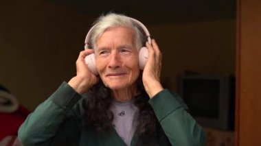 Yaşlı kadın kablosuz kulaklık kullanıyor. Yaşlılar ve teknolojiler. Köy emeklisi, kablosuz kulaklıkla müzik dinliyor. Mutlu emeklilikler, insanlar ve aletler
