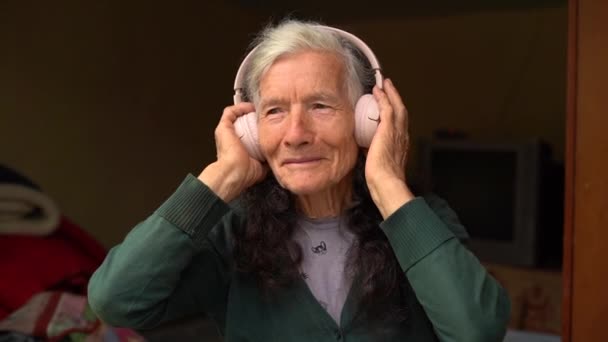 คนแก่และเทคโนโลยี นักเกษียณหมู่บ้านฟังเพลงด้วยหูฟังไร้สาย มีความสุขกับการเกษียณอายุ คนและอุปกรณ์ ผู้หญิงแก่ใช้หูฟังไร้สาย — วีดีโอสต็อก