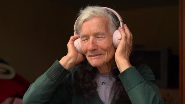 Une femme de la campagne âgée écoute de la musique pour la première fois grâce à un casque sans fil. Vieille dame et la technologie moderne, profiter de la musique avec les yeux fermés — Video
