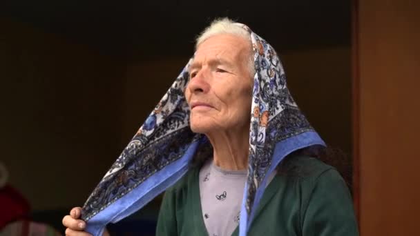 Oude plattelandsvrouw met grijs haar en een gerimpeld gezicht bindt een sjaal over haar hoofd. Authentiek portret van een oudere dame uit Oost-Europa — Stockvideo