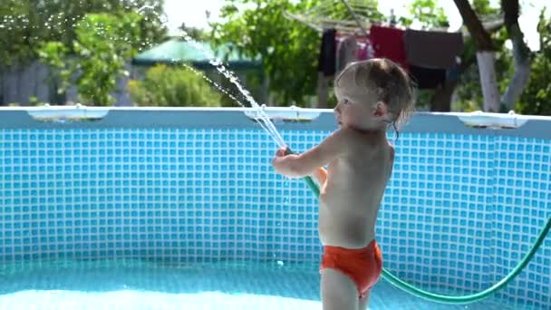 Menino alegre de três anos está brincando com um jato de água de uma mangueira enquanto está em uma piscina inflável. Verão férias e água jogos conceito — Vídeo de Stock