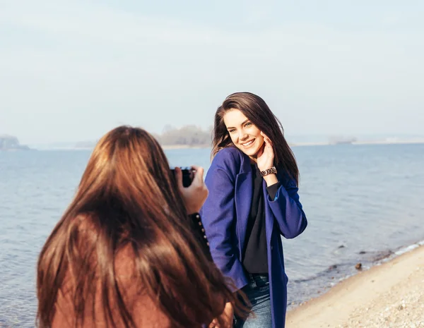 Cheerful young girl in a blue coat posing her girlfriend on the beach — Stock Photo, Image