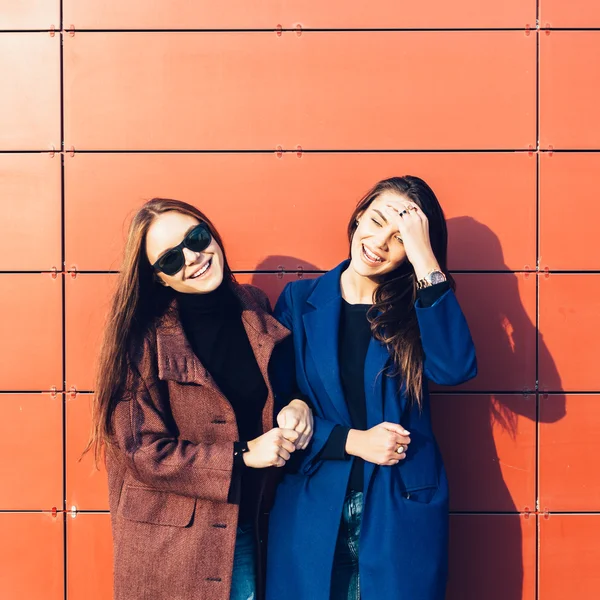 Dos hermosas amigas jóvenes en abrigos posando cerca de la pared roja —  Fotos de Stock