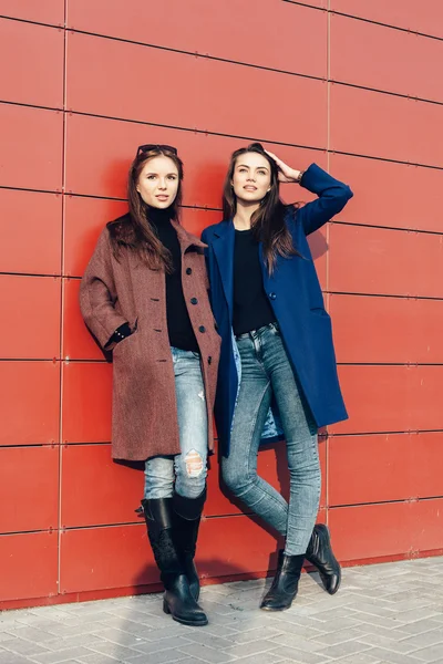 Two beautiful young girl friends in  coats posing near red wall — Stock Photo, Image