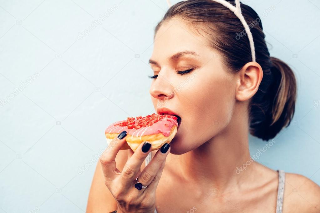 Download - Attractive brunette sexy woman eating tasty donut. Outdoors lifestyle portrait of pretty girl - Stock Image