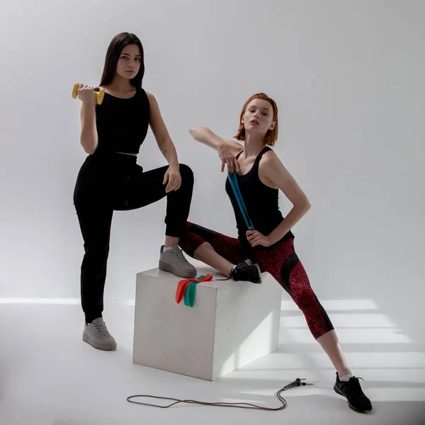 Parlak spor giyimli iki tatlı kız parlak bir odada elastik bantlarla spor yapmaya gidiyorlar.